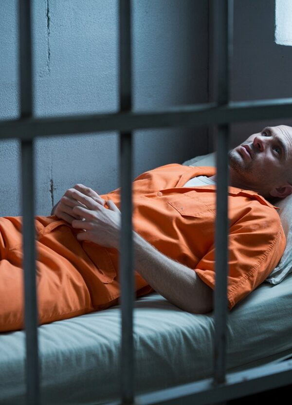 Homem deitado em uma cama dentro de uma cela de prisão, parecendo fraco e abatido.